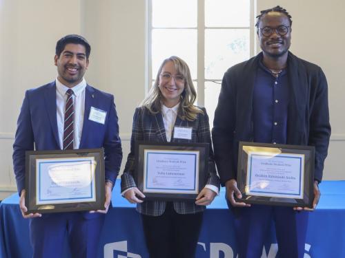 Three people stand in front of a table with a blue table cloth, holding their 2024 Library Graduate Student Prize certificates