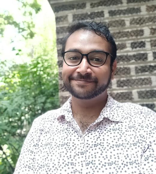 A man in a white shirt and glasses standing in front of a brick wall and greenery