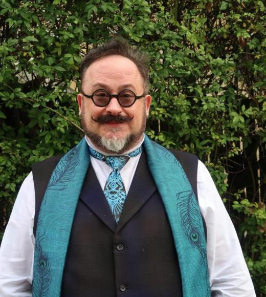 A man wearing round classes, a black vest, and a green necktie and scarf