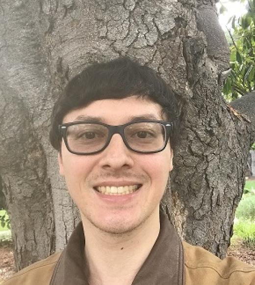Man with glasses in front of tree wearing brown jacket
