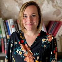 Smiling blonde woman in floral blouse standing before bookshelves and a world map