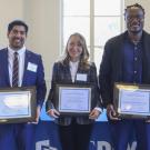 Three people stand in front of a table with a blue table cloth, holding their award certificates