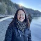 A woman wearing a coat near a body of water and a forest.