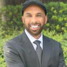 Smiling bearded man in black suit and cap, headshot outdoors against green shrubs