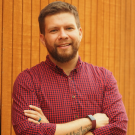 A man in a read shirt with a short beard in front of a wooden wall