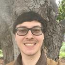 Man with glasses in front of tree wearing brown jacket